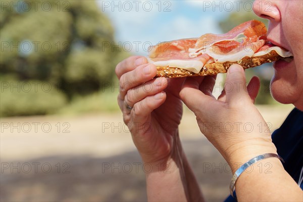 Woman in profile eating a toast of bread with tomato and acorn-fed iberian ham in the countryside with a forest in the background and a blue sky with clouds