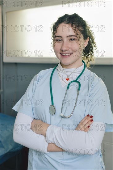 Smiling young female doctor medical nurse wearing white coat and stethoscope around neck standing in private clinic with arms crossed looking at camera. Vertical portrait