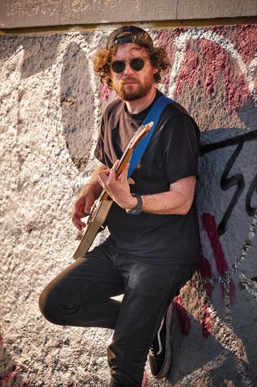 Hipster street musician in black playing electric guitar in the street on sunset leaning on a wall