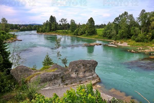 The river bank of the Lech in Lechbruck with a view of a rock formation. In the background is a wooded area with trees and a blue sky with white clouds. The picture has a peaceful and calm mood. Lechbruck