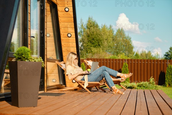 Woman watching movie on laptop while resting in a folding wooden sun lounger on the terrace of log cabin
