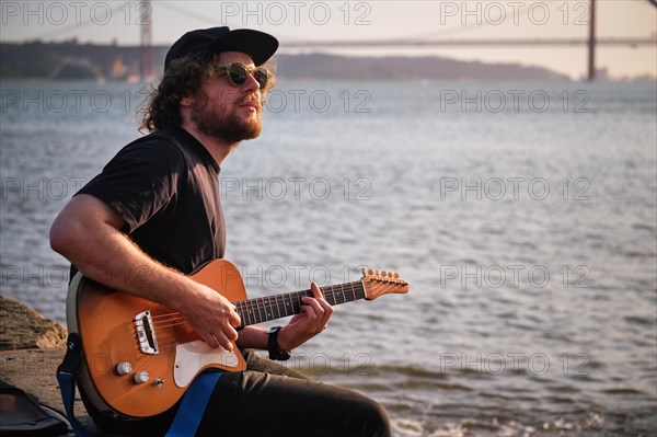 Hipster street musician in black playing electric guitar in the street sitting on pier embankment on sunset in Lisbon