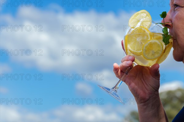 Woman seen in profile drinking a glass of gin and tonic with lemon slices and mint leaves with a blue sky with clouds in the background
