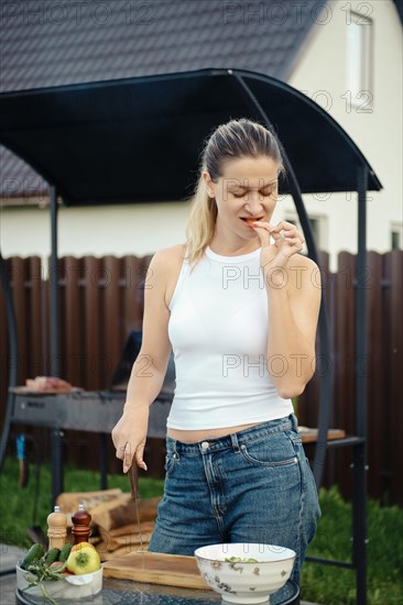 A woman rests her knife on a cutting board and bites a slice of cucumber