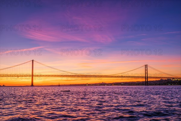 View of 25 de Abril Bridge famous tourist landmark of Lisbon connecting Lisboa and Almada over Tagus river with tourist yacht silhouette at sunset. Lisbon