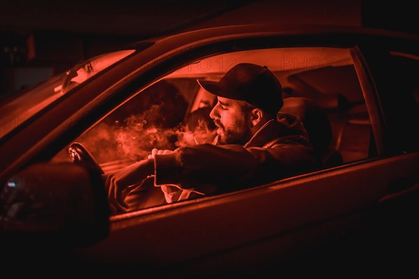 Man in a cap driving car smoking at night in a garage lit with a red light