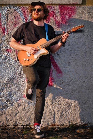 Hipster street musician in black playing electric guitar in the street on sunset leaning on a wall. Lisbon