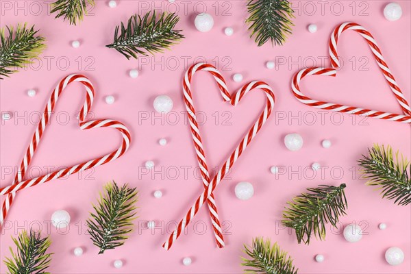 Christmas candy canes forming heart surrounded by white snowball ornaments and fir branches on pink background