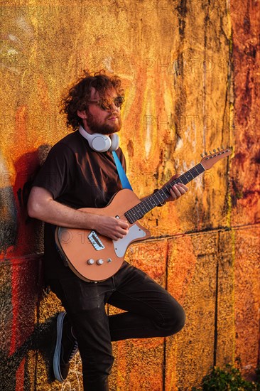 Hipster street musician in black playing electric guitar in the street on sunset leaning on a painted wall