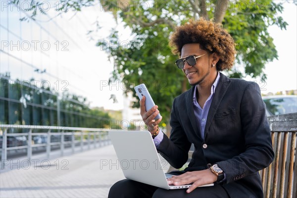 Cool businessman using mobile phone and laptop while working sitting on a urban park bench