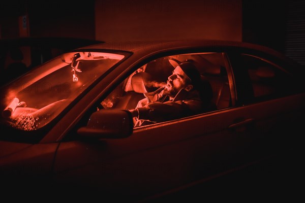 Rapper man in a cap in a car smoking at night in a garage lit with a red light in winter