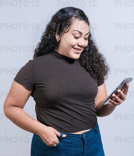 Smiling latin girl using cell phone isolated. Cheerful young woman with curly hair texting on cell phone isolated