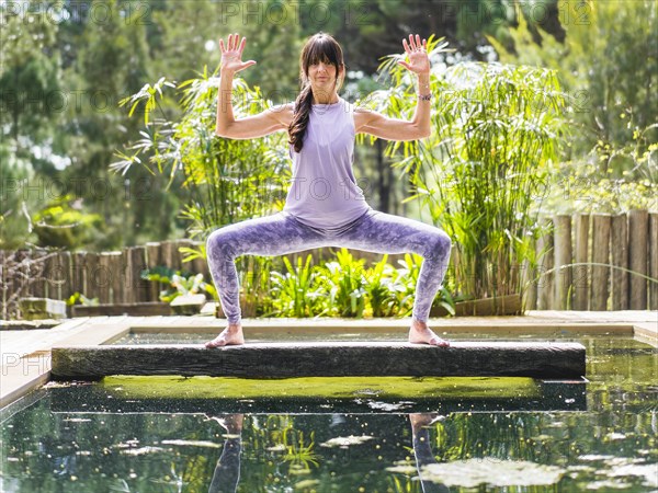 A woman practicing the yoga pose Goddess with Cactus Arms on a tree trunk over a pool