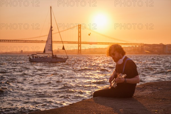 Hipster street musician in black playing electric guitar in the street on sunset on embankment with 25th of April bridge and yacht boat in background. Lisbon