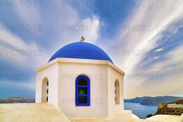 White Greek Orthodox church and its blue dome with cross against dramatic blue sky and clouds