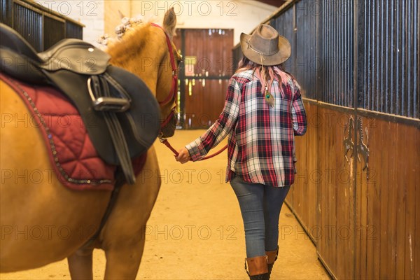 Unrecognizable cowgirl woman strolling with a horse in a stable