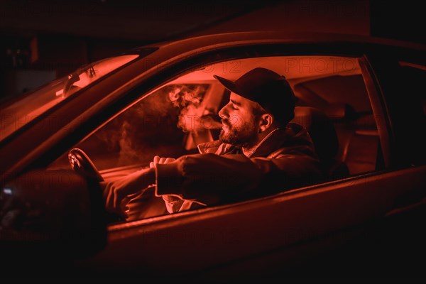 Man in a cap driving car smoking at night in a garage lit with a red light