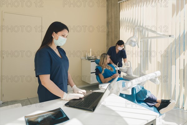 Female dentist typing patient data on laptop computer while patient converses with assistant in dental office chair