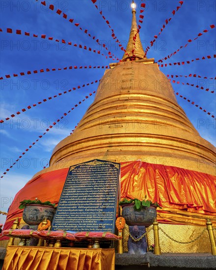 Stone board with with long message in Thai in front of big golden stupa or chedi decorated with festive red flags and textile in sunset sunlight in Wat Saket or Golden Mount temple