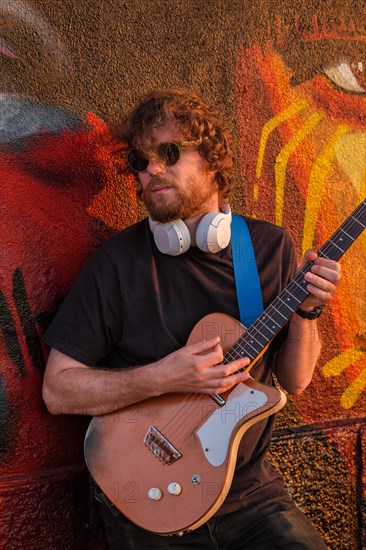 Hipster street musician in black playing electric guitar in the street on sunset leaning on a painted wall