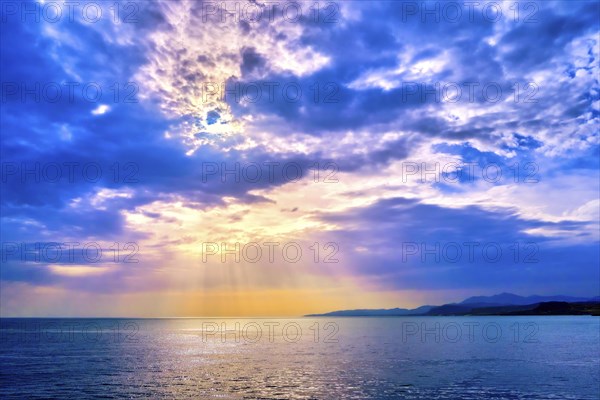 Beautiful seascape with colorful clouds and bright sun rays cast over calm sea waters and hazy distant shores. Sunrise or sunset in Mediterranean sea