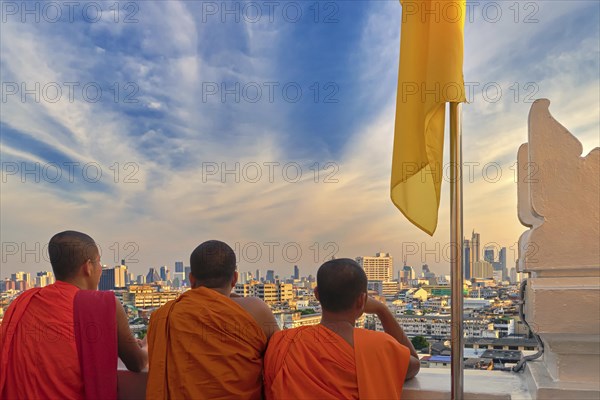 Three male Buddhist monks stand by white parapet wall of Wat Saket or Golden Mount temple and look at skyline of Bangkok