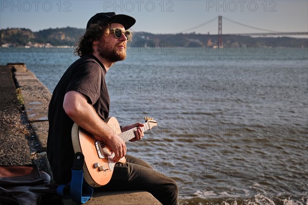 Hipster street musician in black playing electric guitar in the street sitting on pier embankment on sunset in Lisbon