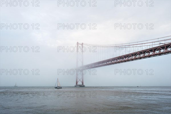 View of 25 de Abril Bridge famous tourist landmark of Lisbon connecting Lisboa and Almada in heavy fog mist wtih yacht boats passing under. Lisbon