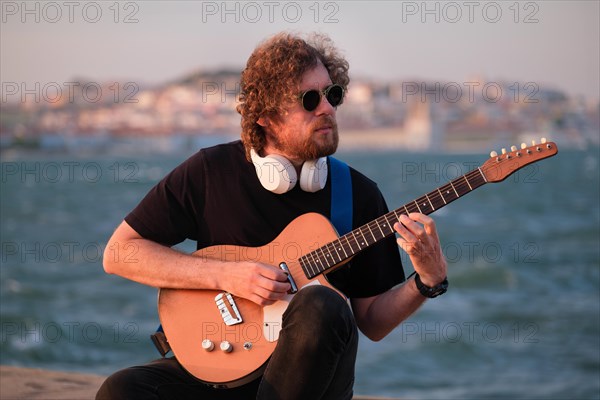 Hipster street musician in black playing electric guitar in the street on sunset on embankment