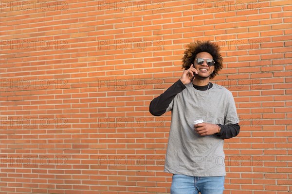 Horizontal photo with copy space of a Mixed-race man using mobile and holding a take away coffee next to a brick wall