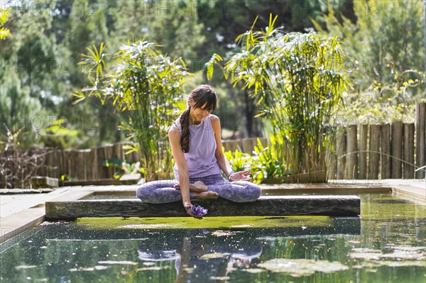 A woman holding a glass purple lotus while sitting in lotus pose on a tree trunk over the water