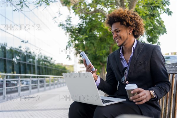 Cool african american businessman working with laptop and mobile sitting on the street in the morning