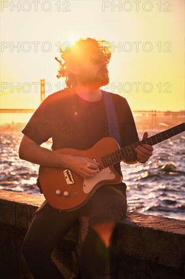 Hipster street musician in black playing electric guitar in the street on sunset on embankment with 25th of April bridge in background. Lisbon