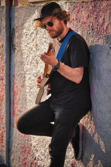 Hipster street musician in black playing electric guitar in the street on sunset leaning on a wall. Lisbon