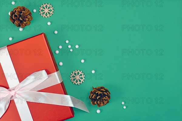 Christmas gift box with ribbon surrounded by seasonal pine cones and wooden snowflake ornaments in corner of green background with empty copy space