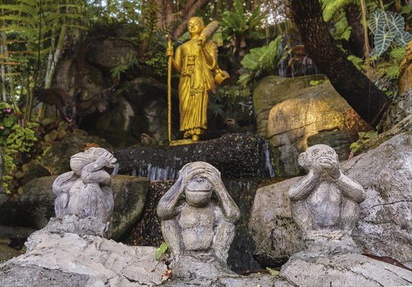 Buddhism symbols: gilded statue of Buddha as walking monk with Naga walking stick and umbrella and three wise monkeys statues sitting in foreground. Religion