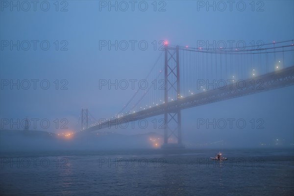 View of 25 de Abril Bridge famous tourist landmark of Lisbon connecting Lisboa and Almada in heavy fog mist wtih yacht boats passing under. Lisbon