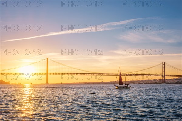 View of 25 de Abril Bridge famous tourist landmark of Lisbon connecting Lisboa and Almada over Tagus river with tourist yacht silhouette at sunset. Lisbon