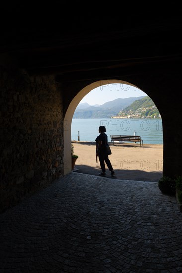 Woman Walking in an Old Beautiful Street Tunnel with Arch from Brusino Arsizio on the Waterfront in a Sunny Summer Day and with Lake Lugano and Mountain View over Morcote