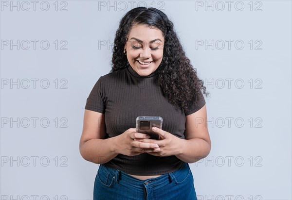 Cheerful young woman with curly hair texting on cell phone isolated. Smiling latin girl using cell phone isolated