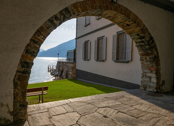 Beautiful Arch to a Park with a House on the Waterfront to Lake Lugano with Mountain in a Sunny Summer Day in Bissone