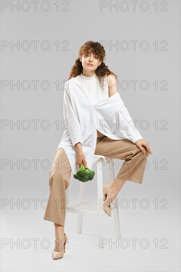 Beautiful woman with broccoli sits on tall chair in grey studio