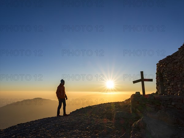 Mountaineer standing at sunset next to summit chapel Timios Stavros and summit cross of Psiloritis