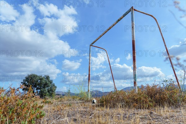 Old rusty soccer goal in an overgrown field with a broken soccer ball in front of it