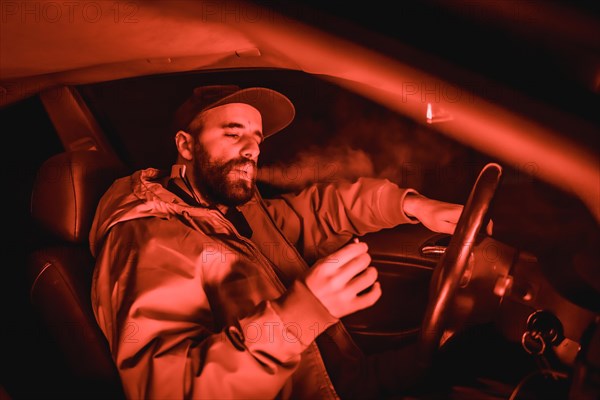 Rapper man in a cap in a car smoking at night in a garage lit with a red light in winter