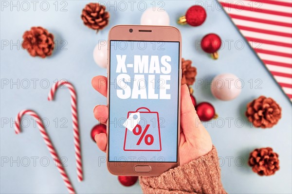 Concept for Christmas seasonal online shopping and sales with hand holding cell phone with 'X-Mas Sale' sign in front of desk with seasonal decorations