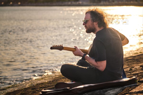 Hipster street musician in black playing electric guitar in the street sitting on pier embankment on sunset