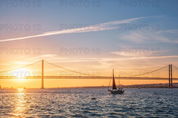 View of 25 de Abril Bridge famous tourist landmark of Lisbon connecting Lisboa and Almada over Tagus river with tourist yacht silhouette at sunset. Lisbon