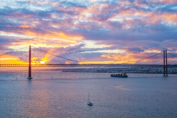 View of 25 de Abril Bridge famous tourist landmark of Lisbon over Tagus river with tourist yacht boats and cargo container ship on sunset. Lisbon