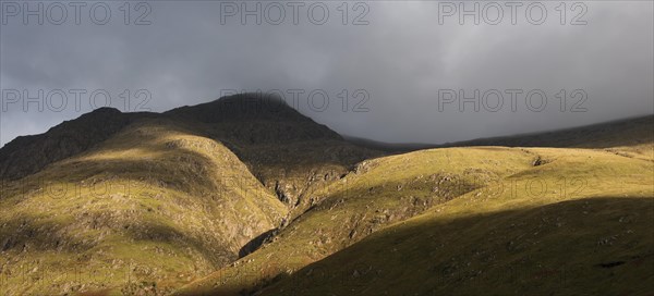 Rain cloud forming thick fog descending from steep mountainside bading in evening light at sunset in Glen Coe